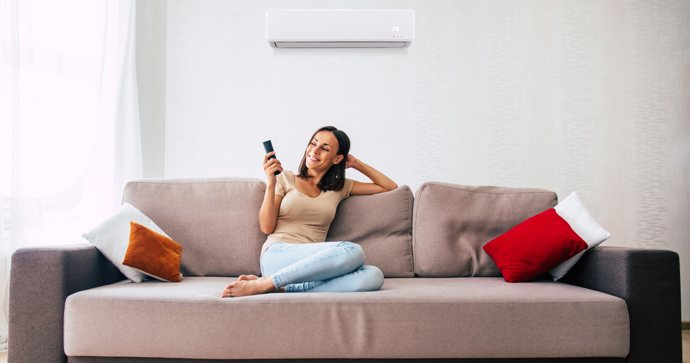dark haired woman in blue jeans sat on sofa smiling using air conditioning remote control